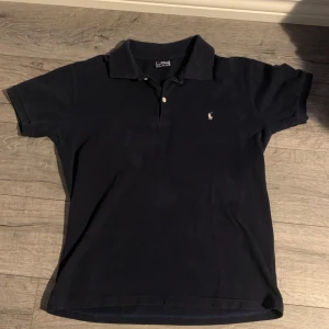 Svart pikétröja från Ralph Lauren  - Klassisk svart pikétröja från Ralph Lauren i storlek M. Tröjan har korta ärmar, krage och knäppning framtill samt den ikoniska broderade loggan på bröstet. Tillverkad i mjuk bomull som är skön att bära hela dagen.
