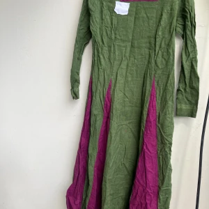 Grön och lila långklänning i linne - Unik långklänning i grönt linne med lila paneler och långärmad design. Klänningen har en rak passform och färgblockad look som sticker ut. Perfekt för dig som gillar färg och vill ha något annorlunda i garderoben. Den är hel och ren. Maskinsydd.  Vidd ca 80cm (över brösten), längd ca 115cm. Lite skrynklig men det är allt jag kan hitta att anmärka på. Även om den inte är i nyskick.