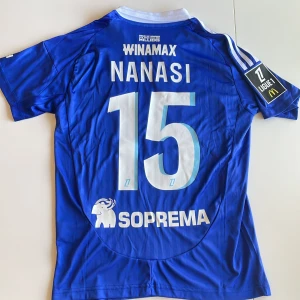 Strasbourg Nanasi #15 matchtröja Adidas - Blå officiell RC Strasbourg fotbollströja replika från Adidas med Nanasi och nummer 15 på ryggen. Tröjan har vit krage, vita detaljer och sponsorloggor. Kortärmad modell i lätt funktionsmaterial, perfekt för match eller träning. Ligue 1-märke på ärmen. Endast handtvätt!