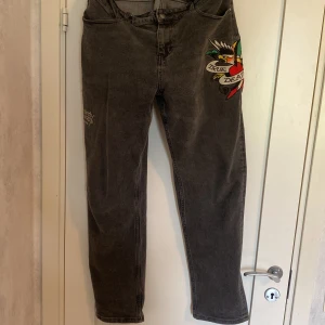 Grå jeans från Ed Hardy med brodyr - Snygga grå jeans från Ed Hardy med stor broderad logga på bakfickorna och färgglad tattoo-inspirerad brodyr framtill. Klassisk femficksmodell i jeansmaterial med normal passform och raka ben. Perfekta för dig som vill sticka ut med coola detaljer.