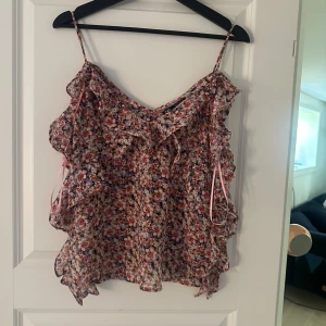Blommig volangtop från Zara - Supersöt blommig linnetopp från Zara med tunna axelband och volangdetaljer. Toppen har en mix av rosa, lila och vita blommor på en ljus botten och en liten rosett framtill. Perfekt för varma dagar och ger en romantisk vibe.