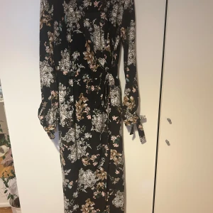 Svart blommig långklänning med knyt Nelly - Supersnygg svart långklänning med blommigt mönster i beige, vitt och rosa. Klänningen har långa ärmar, v-ringning och knytband i midjan för en snygg siluett. Perfekt för dig som gillar romantiska vibes och vill sticka ut lite extra.