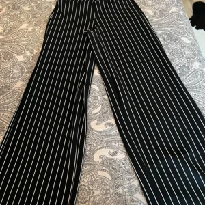 Svarta randiga wide byxor - Snygga svarta byxor med vita vertikala ränder och breda ben. De har en hög midja och loose passform, perfekta för en trendig och avslappnad stil. Materialet känns mjukt och följsamt.