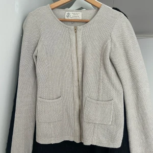 Beige stickad cardigan jacka Newhouse - Beige stickad cardigan jacka från Newhouse med rund halsringning, två framfickor och guldfärgad dragkedja. Snyggt strukturerad yta och klassisk passform som funkar till många olika looks.