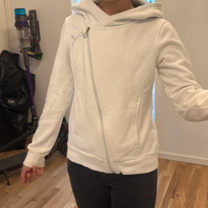 Vit hoodiejacka från Puma med dragkedja - Säljer en vit hoodiejacka från Puma med sned dragkedja och broderad logga på bröstet. Jackan har huva, långa ärmar och är gjord i mjukt bomullsmaterial. Perfekt för dig som gillar sportig och clean stil.