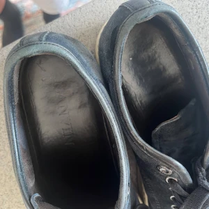 Svarta sneakers från Lanvin i mocka - Snygga svarta sneakers från Lanvin med ovandel i mocka och lackad tå. Skorna har svarta snören, silverfärgade öljetter och vit sula med Lanvin-logga undertill. Perfekta för dig som gillar stilrena och exklusiva sneakers med en twist.