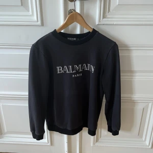 Svart sweatshirt från Balmain Paris - Svart sweatshirt från Balmain Paris med vit logga framtill. Klassisk rund halsringning och ribbade muddar vid ärmslut och nederkant. Tillverkad i mjuk bomull för en skön och avslappnad look. Perfekt för dig som gillar stilrena designer och streetwear vibes.