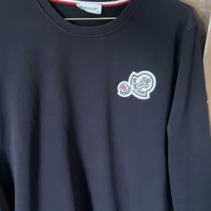 Svart långärmad tröja från Moncler - Svart långärmad tröja från Moncler med rund halsringning och snyggt broderat logomärke på bröstet. Tröjan har en stilren och enkel design som passar perfekt till jeans eller chinos. Materialet känns mjukt och bekvämt mot huden.