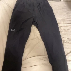 Svarta träningsbyxor från Under Armour - Svarta träningsbyxor från Under Armour i storlek M. Byxorna har en slim passform, elastisk midja med snörning och är tillverkade i ett syntetmaterial som andas. Perfekta för gymmet eller löprundan, med diskret logga på benet.