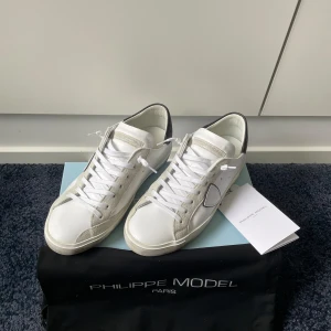 Vita Philippe Model sneakers - Snygga vita sneakers från Philippe Model med detaljer i grå mocka och svart hälparti. Skorna har klassisk snörning, rund tå och platt sula. Tillverkade i skinn och mocka för en lyxig känsla. Perfekta för dig som gillar stilrena och trendiga sneakers.                                                              Köpta på merchsweden