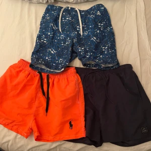 Bad byxor från Volcom, Polo & Rip Curl  - Tre par badshorts: ett blått mönstrat från Volcom med vit snörning, ett knallorange från Polo Ralph Lauren med svart logga och svart snöre, samt ett mörkblått från Rip Curl med diskret logga. Perfekta för stranden eller poolen! De blåa är i storlek 170 och orange i L och de mörkblå är i M. Om man köper alla tillsammans blir de 550kr annars 200kr/st
