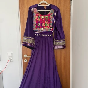 Afgansk lila klänning - Unik afghansk långklänning i lila sammet med breda ärmar och färgglatt broderat bröstparti i rött, grönt och blått. Klänningen har guldiga detaljer, spegelprydda dekorationer och mynt längs midjan. Perfekt för dig som vill sticka ut med traditionell och färgstark stil.