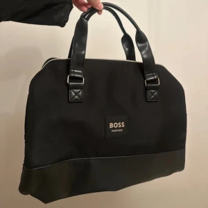 Svart handväska från BOSS Parfums - Stilren svart handväska från BOSS Parfums med två rejäla handtag och detaljer i skinnimitation. Väskan har en klassisk form, dragkedja upptill och diskret logga framtill. Perfekt för dig som vill ha en snygg och praktisk väska med plats för allt du behöver.