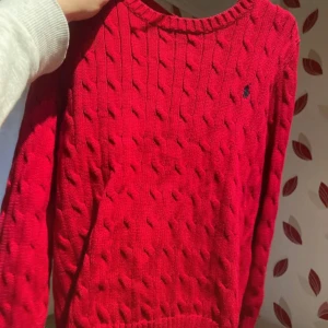 Röd kabelstickad tröja Polo Ralph Lauren - Kabelstickad röd tröja från Polo Ralph Lauren med klassisk rund halsringning och broderad marinblå logga på bröstet. Tröjan har lång ärm och ribbade muddar, perfekt för lager-på-lager under kyliga dagar.