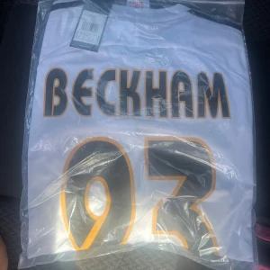 Vit Real Madrid Beckham tröja Adidas - Säljer en vit Real Madrid fotbollströja från Adidas med Beckham och nummer 23 i svart och guld på ryggen. Tröjan har klassisk passform och är tillverkad i lätt och ventilerande material, perfekt för dig som älskar fotboll och vill ha en ikonisk look.