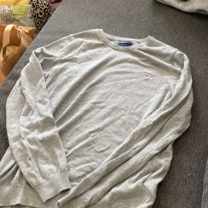 Grå stickad tröja från GANT XS - Säljer en klassisk grå stickad tröja från GANT i storlek XS. Tröjan har rund halsringning, långa ärmar och en broderad GANT-logga med sköld på bröstet. Perfekt för lager-på-lager och enkel att matcha med jeans eller kjol.