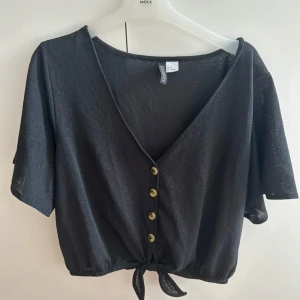 Svart croppad blus med knyt och knappar - Svart croppad blus med v-ringning, korta ärmar och snygga knappar framtill. Nedtill finns ett knyt som ger en cool och avslappnad vibe. Perfekt till jeans eller kjol för en trendig look