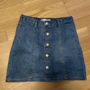 Levi's jeanskjol med knappar fram - Snygg blå jeanskjol från Levi's med hög midja och raka linjer. Kjolen har fem silverfärgade knappar framtill och klassiska bälteshällor. Perfekt för dig som gillar en tidlös denimlook.