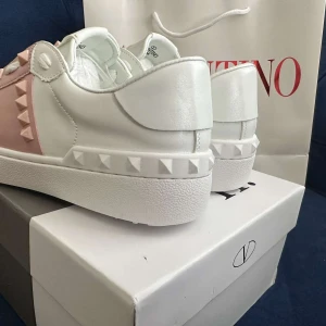 Valentino Rockstud sneakers vit/rosa - Snygga Valentino Rockstud sneakers i vitt skinn med ljusrosa detaljer och ikoniska nitar. Klassisk rund tå, platt sula och snörning framtill. Perfekta för dig som vill ha en lyxig och trendig look med en touch av rosa.