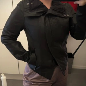 Svart skinnjacka från Lindex - En svart skinnjacka från Lindex med dragkedja framtill och en hög krage. Jackan har långa ärmar och detaljer som knappar vid kragen.