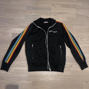 Palm Angels svart vindjacka med regnbågsränder - Svart vindjacka från Palm Angels med vita dragkedjor och regnbågsfärgade ränder längs ärmarna. Jackan har ståkrage, två fickor med dragkedja och logga broderad på bröstet. 