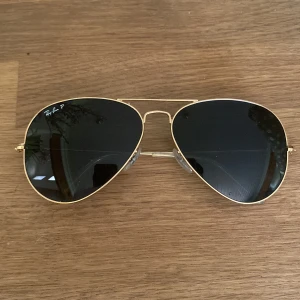 Ray-Ban Aviator solglasögon guld - Snygga Ray-Ban Aviator solglasögon med tunna guldfärgade metallbågar och mörka, polariserade linser. Klassisk pilotform som ger en tidlös vibe. Perfekta för att lyfta din stil på sommaren.