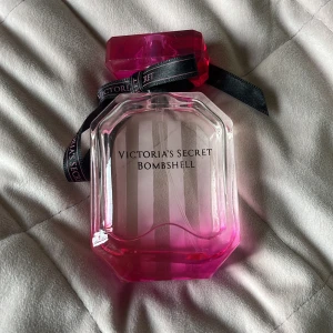 Victoria's Secret Bombshell parfym - Säljer en ikonisk Victoria's Secret Bombshell parfym i en snygg, åttakantig glasflaska med rosa toning och ett stort rosa lock. Flaskan har svarta band med logga och vertikala ränder. Perfekt för dig som gillar fräscha och feminina dofter.