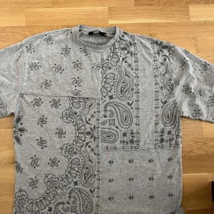 Grå paisleymönstrad t-shirt från Jack & Jones - Säljer en grå t-shirt med svart paisleymönster och patchwork-känsla. T-shirten har korta ärmar, rund hals och är gjord i mjuk bomull. Perfekt för dig som gillar streetstyle och unika prints.