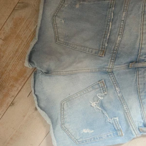 Ljusblå slitna jeansshorts Zara  - Säljer ett par ljusblå jeansshorts med slitna detaljer och fransiga kanter. Shortsen har klassisk femficksdesign, dragkedja och knapp framtill. Perfekt för dig som gillar en edgy och avslappnad stil. 