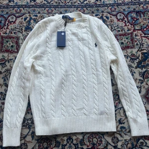 Vit kabelstickad tröja Polo Ralph Lauren - Vit kabelstickad tröja från Polo Ralph Lauren med klassisk rund halsringning och broderad marinblå logga på bröstet. Tröjan har ribbade muddar och är långärmad, perfekt för lager-på-lager under kyliga dagar.