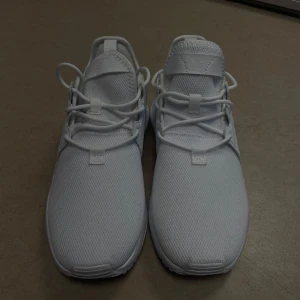 Helvita Adidas sneakers mesh - Snygga helvita Adidas sneakers med klassiska tre ränder på sidan. Ovandelen är i ventilerande mesh och skon har snörning samt platt sula. Perfekta för en clean och sportig look, med diskret Adidas-logga. Helt oanvända