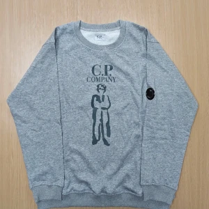 Grå sweatshirt från C.P. Company - Snygg grå sweatshirt från C.P. Company med tryck framtill och klassisk rund hals. Tröjan har ribbade muddar och en ikonisk svart detalj på ärmen. Perfekt för dig som gillar streetwear och vill ha en stilren look.