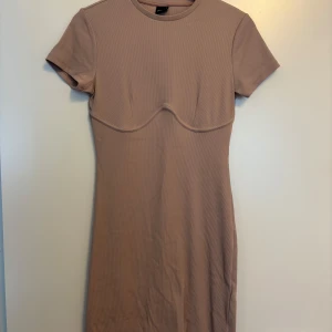 Beige ribbad kortklänning Gina Tricot - Supersnygg ljusrosa färg ribbad kortklänning från Gina Tricot. Klänningen har korta ärmar, rund halsringning och markerad söm under bysten för en cool detalj. Perfekt för dig som gillar en enkel men trendig look.