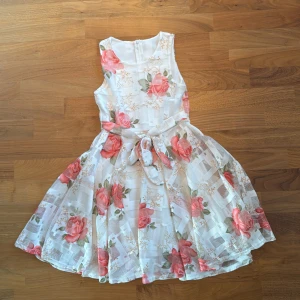 Vit blommig kortklänning med rosett - Superfin vit kortklänning med stora rosa blommor och gröna blad. Klänningen är ärmlös, har rund halsringning och en söt knytrosett i midjan. Tillverkad i 100% polyester och har dragkedja i ryggen. Perfekt för somriga dagar eller speciella tillfällen.