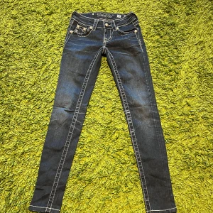 Miss Me skinny jeans med detaljer - Mörkblå skinny jeans från Miss Me med vita kontrastsömmar och snygga broderade detaljer samt paljetter på bakfickorna. Jeansmodellen är tight och har dekorativa nitar och logga på bakfickan. Perfekt för dig som gillar statement-jeans.