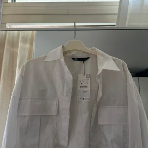 Vit croppad skjorta från Zara i bomull - Vit overshirt från Zara i bomull med klassisk krage och stora bröstfickor. Skjortan har en rak passform och knäppning framtill. Gul missfärgning som bör gå bort med en ordentlig tvätt. Prislapp kvar 