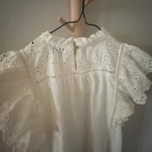 Vit blus med volang från Holly Whyte - Superfin vit blus från Holly Whyte med broderade detaljer och volangärmar. Blusen har romantisk spets, rynkade partier och en liten ståkrage med volang. Perfekt för dig som gillar en söt och luftig stil.