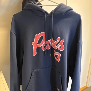 Jordan Hofor med loggan Paris 23 - Mörkblå hoodie med huva och snörning, stor ficka framtill och tryck i rött och vitt med texten 'Paris 23' på bröstet. Perfekt för dig som gillar streetstyle och vill ha en skön och avslappnad look.