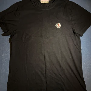 Svart Moncler t-shirt med logga - Svart t-shirt från Moncler i mjuk bomull. Snygg och enkel design som passar till allt. Röd, vit och blå randig detalj på insidan av kragen ger en extra touch.
