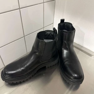 Svarta chelsea boots i skinn - Stilrena svarta chelsea boots med chunky sula och elastiska paneler på sidorna. Tillverkade i slätt skinn och har en rund tå samt grovt mönstrad yttersula för extra grepp. Perfekta för dig som vill ha en klassisk men edgy look.