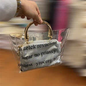 Transparent handväska med glitterdetaljer - Säljer en unik transparent handväska med guldiga handtag och en innerdel i glittrigt silvertyg. Väskan har tryckt text på framsidan och kedjedetaljer i guld. Perfekt för dig som vill sticka ut och visa din stil.