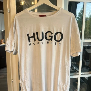 Vit t-shirt från Hugo Boss - Vit t-shirt från Hugo Boss med stor svart logga framtill. Klassisk rund hals och korta ärmar. Tillverkad i mjuk bomull som känns skön mot huden. Perfekt för dig som gillar stilrena och enkla plagg med tydlig branding.
