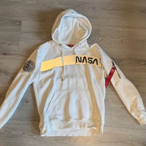 Vit Alpha industries NASA hoodie med patch och dragkedja - Snygg vit hoodie från alpha industries med NASA-tryck och silvrig detalj på bröstet. Tröjan har huva med snörning, känguruficka och en alphas ordinal patch på ena ärmen. På ärmen finns även en liten ficka med dragkedja och röd tag. Perfekt för dig som gillar rymd och streetstyle. Pris 399 priset är diskuterbart högst bjudande!