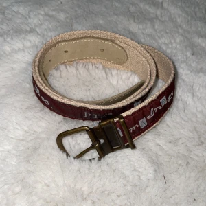 Mörkbrunt bälte från PTA med text - Snyggt mörkbrunt bälte från PTA med texten 'mid Summer madness' tryckt längs hela bandet. Bältet har beige detaljer och ett spänne i metall. Perfekt accessoar för att lyfta din outfit med en unik touch. Ca 94 cm långt.