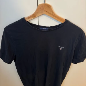 Svart t-shirt från GANT i bomull - En svart t-shirt från GANT med klassisk passform och rund hals. T-shirten har GANT-logga broderad på bröstet och är tillverkad i mjuk bomull. Perfekt basic-plagg med korta ärmar och stilren look. Nypris ligger på 599kr