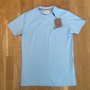 Ljusblå Burberry t-shirt i bomull - Snygg ljusblå t-shirt från Burberry med klassisk rund hals och korta ärmar. T-shirten har ett broderat Burberry-emblem i guld på bröstet och ikoniskt rutigt band på insidan av kragen. Perfekt för en clean och stilren look. Modellen passar en som är mellan 170 cm till en som är 180 cm! Sjukt bekväm inför sommaren! 