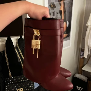 Vinröda boots med gulddetalj - Snygga vinröda boots i skinn med rund tå och cool guldfärgad låsdetalj framtill. Skorna har en stilren design och är perfekta för dig som vill sticka ut med en unik accessoar. Passar till både jeans och kjol för en trendig look.