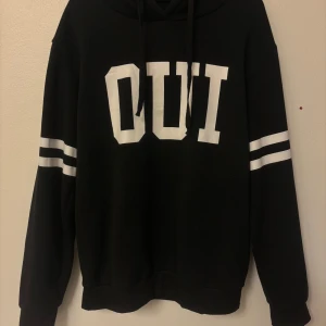 Svart hoodie med vita ränder och tryck - Svart hoodie med stora vita bokstäver 'OUI' framtill och dubbla vita ränder på båda ärmarna. Klassisk huva med snörning och långärmad modell. Perfekt för en chill och sportig look.