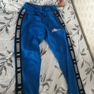 Blå mjukisbyxor från Hoodrich - Snygga blå mjukisbyxor från Hoodrich med svarta sidor och vit logga längs benen. Byxorna har resår i midjan med snörning och är tillverkade i ett mjukt syntetmaterial. Perfekta för en chill och sportig stil.