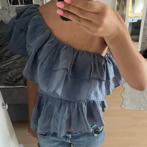 Blå volangig oneshoulder topp - Supersöt blå topp med volanger och oneshoulder-skärning. Luftig och lätt, perfekt för varma dagar. Sitter snyggt med resår i midjan och har flera lager volanger som ger en cool och trendig look.